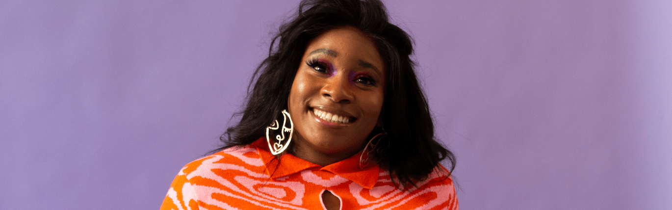 A black women in a pink and orange jumper sits smiling against a purple backdrop with gold earrings.