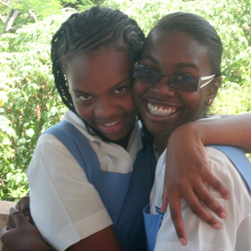 Two school girls where blue pinafores and white shirts hug each other.