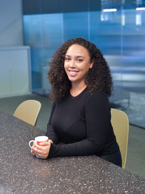 Black woman sits in a conference room, holding a mug in her hands. She smiles at the camera.