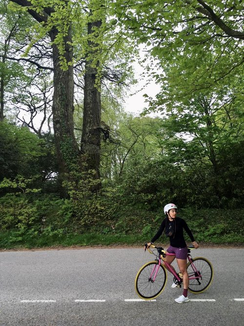 Woman on a bike wearing a helmet and cycling shorts. She's on an empty country road with a forest in the background.