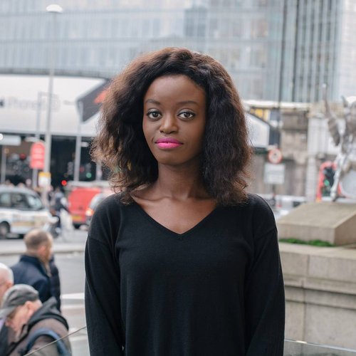 Young black woman stands outside. Behind her is a high rise glass building, and a busy London street.