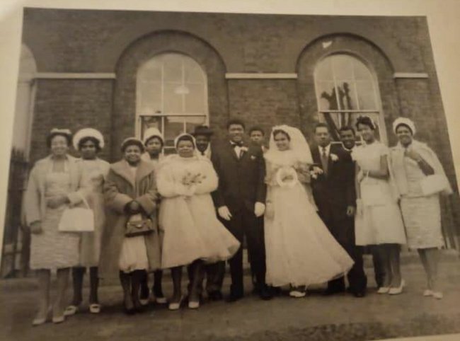 Old wedding photo of thirteen black middle aged people, eight women and five men dressed in formal church clothes stood outside of a building with large arched windows.