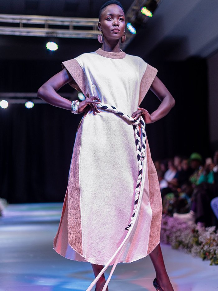 Black model wearing a tunic in beige tones with a rope belt stands with her hands on her hips.
