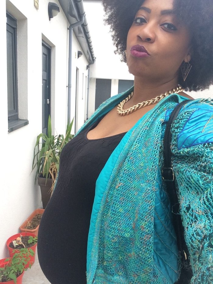 Close up of a pregnant black woman posing for a selfie. She pouts wearing dark purple lipstick, a turquoise knitted top and black blouse.