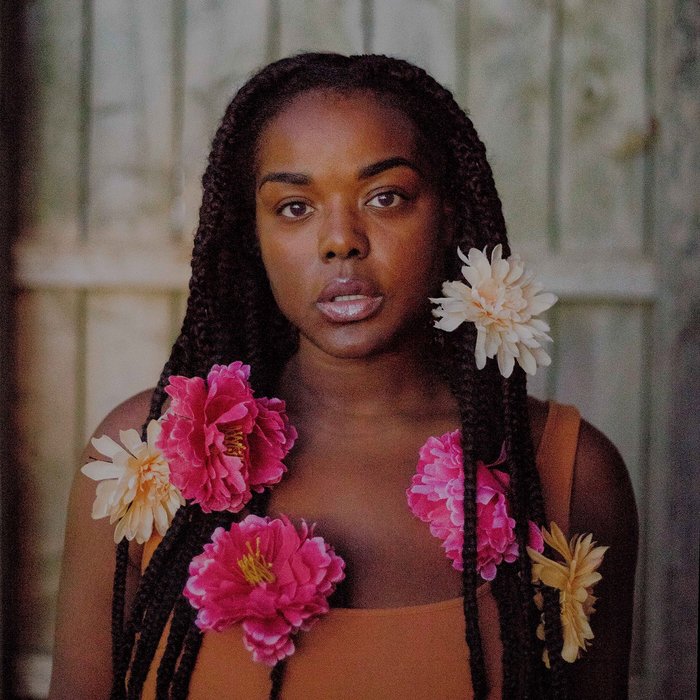 Black woman with bright flowers woven in her long dark braids looks into the camera. Her face is expressionless and her lips are slightly parted.