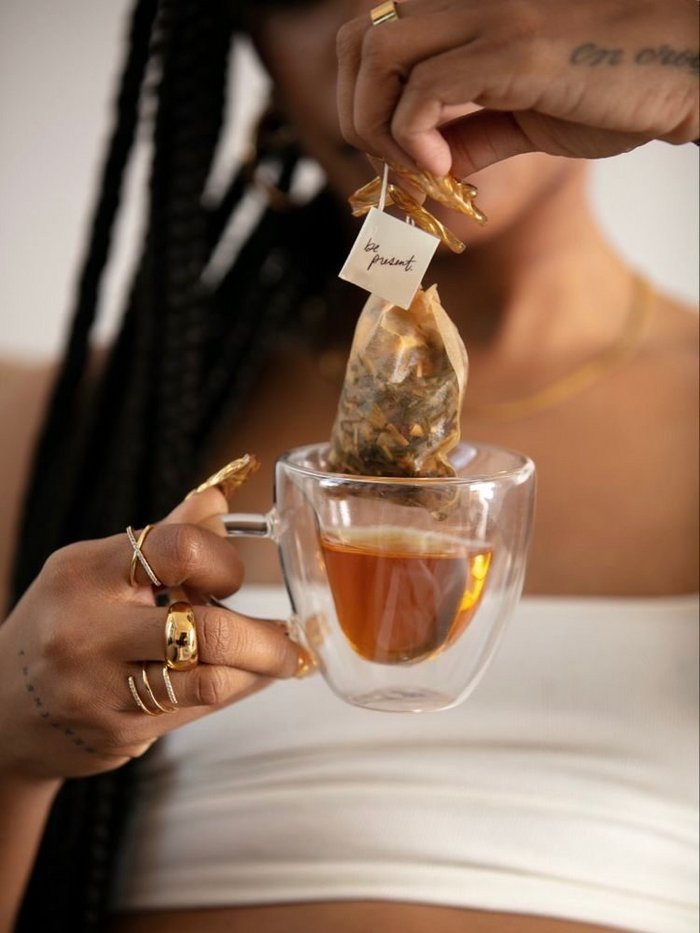 A black woman is dipping a herbal tea bag in a glass cup, she has gold rings on her fingers.