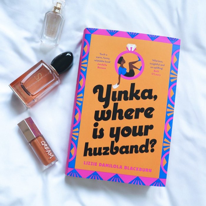 Copy of 'Yinka, Where Is Your Huzband?' laying on a white sheet with a perfume bottle and lip gloss tube next to it.