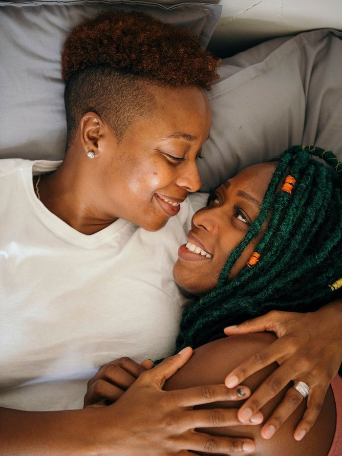 Two black lesbian women are lying in bed together smiling and staring at one another.