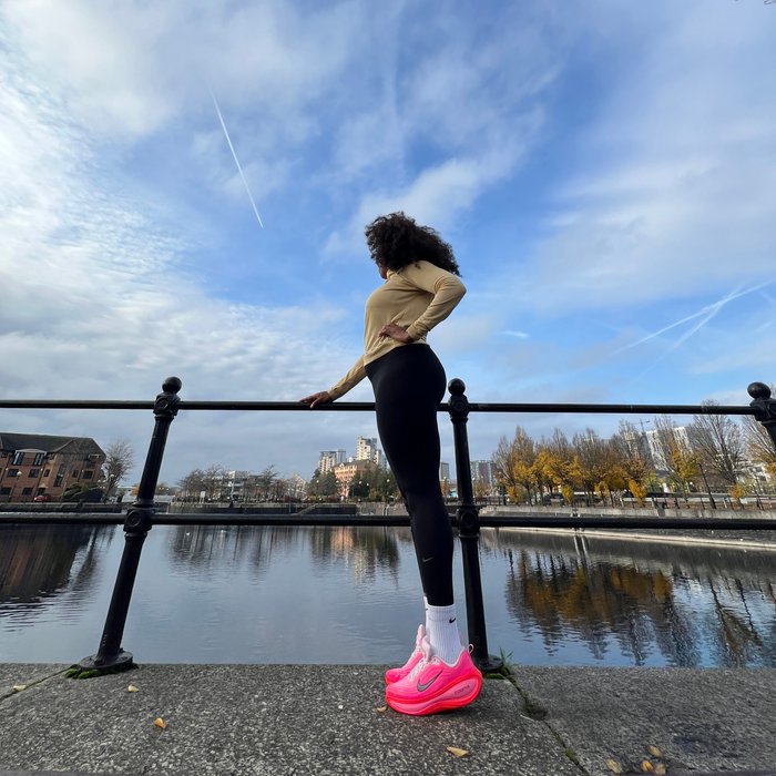 Black woman wearing workout leggings and a long-sleeved workout top stands next to a lake. She rests her hands on the railing and looks out towards the lake. The Sky is blue with wispy clouds and she looks away from the camera, her face obscured by a large curly afro.