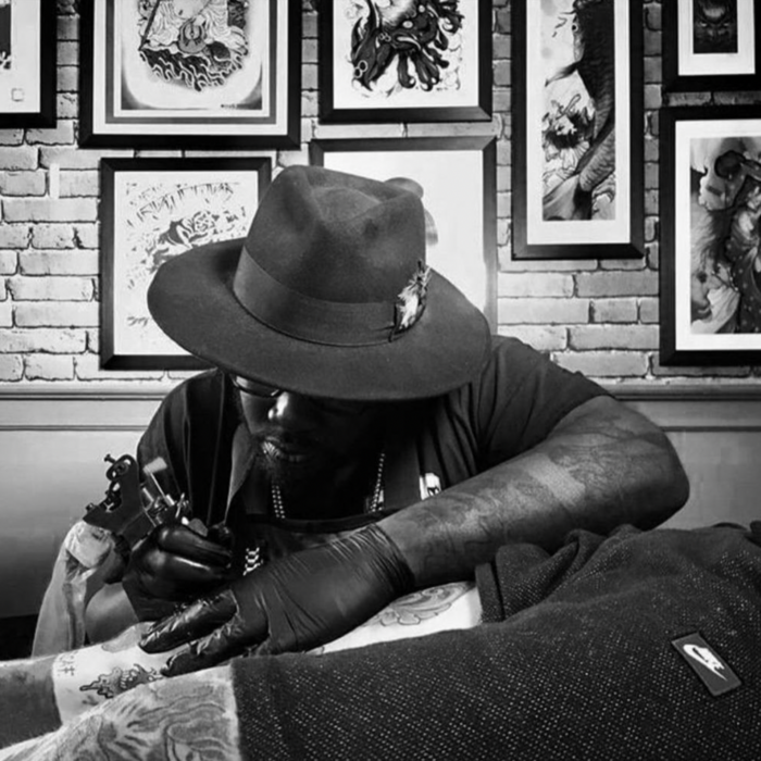 Black and white photograph of a black tattoo artist wearing a wide-brimmed hat, bent over a client tattooing a client's knee.