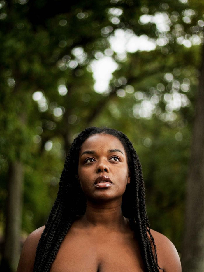 A half body shot of a black woman looking up, she's wearing braids and the background is full of trees.