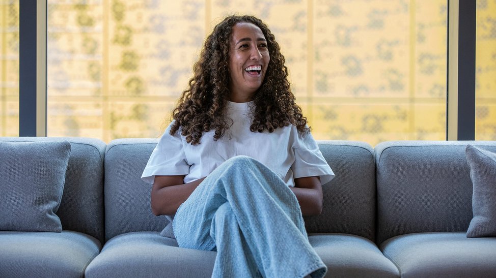 Black woman with long curly hair sits on a sofa, laughing and looking relaxed.