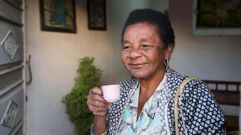 Older black woman holding a small teacup and smiling into the distance.