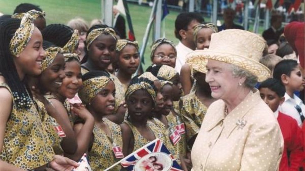 The Queen in a lemon yellow skirt suit with matching hat, smiling at a group of black children in matching print clothing waving Union Jacks.