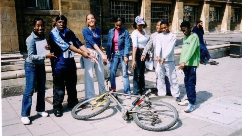 Group of eight young people stand in a semi-circle pointing at a bike that is laying on the floor.