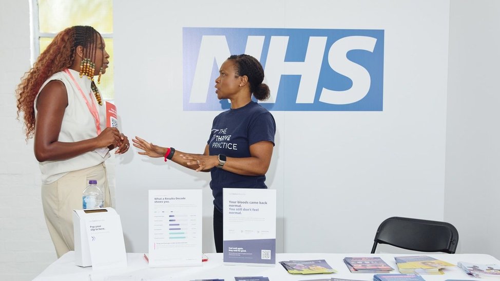 Two black women speaking in front of a wall with the NHS logo.