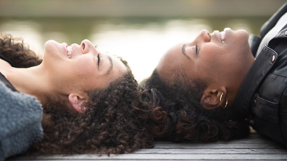 Two black women lie on a bench in front of a lake. They are head to head.