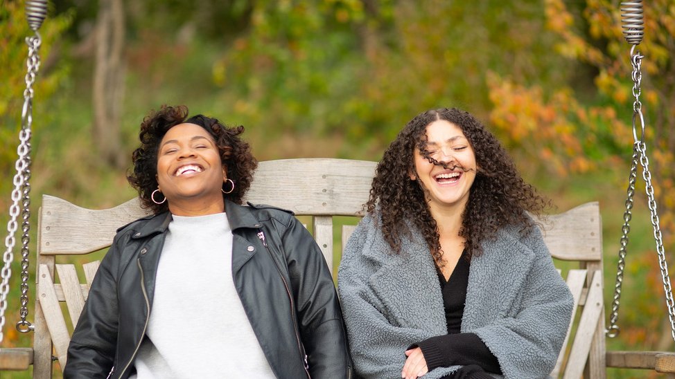 Monique and Yemisi sit side by side on a swinging bench, laughing.