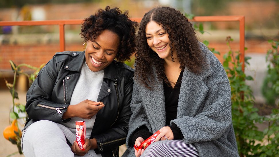 Two black women sit side by side, laughing with bags of Maltesers in their hands.