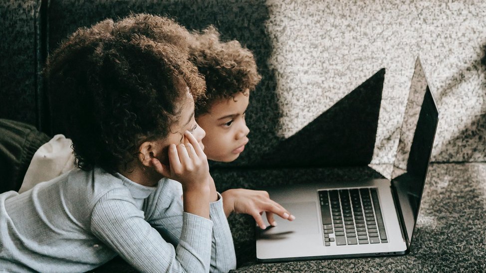 Two young black children lay on their bellies on a dark-coloured sofa. In front of them is an open laptop.